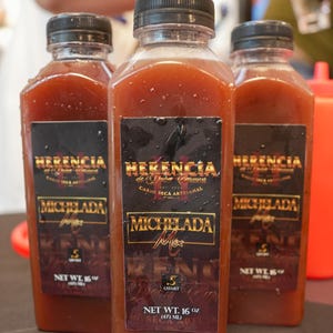 May include: Three square bottles of a reddish-brown liquid, each with a black cap. The labels read "HERENCIA" and "MICHELADA", with additional text. Each bottle contains 16 fl oz (473 ml) of product.