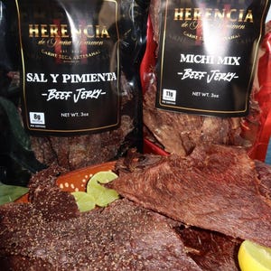 Puede incluir: Dos bolsas de carne seca con etiquetas que dicen "HERENCIA de Doña Carmen" y "SAL Y PIMIENTA - Beef Jerky" y "MICHI MIX - Beef Jerky". La carne seca en rodajas se exhibe en un plato con gajos de lima. La carne seca es de color marrón oscuro.