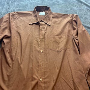 May include: A long-sleeved brown button-down shirt with a single pocket on the left side. The shirt has a white button placket and white buttons.