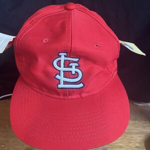 May include: A red baseball cap with a white and blue St. Louis Cardinals logo. The cap has a curved brim and a white tag attached to the side.