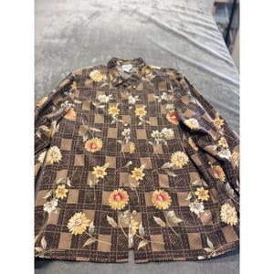 May include: A long-sleeved button-down shirt with a brown plaid pattern and floral accents. The shirt features yellow, orange, and white flowers. The shirt is made of a lightweight material.