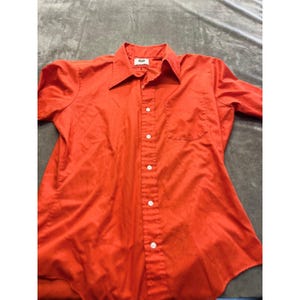 May include: A bright orange button-down shirt with short sleeves. The shirt has a pointed collar, a chest pocket, and white buttons down the front. The shirt is made of a smooth, possibly synthetic fabric.