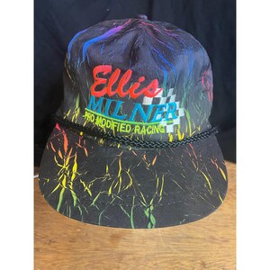May include: Black baseball cap with colorful abstract design and embroidered text "Ellis Milner Pro Modified Racing". The cap has a black rope detail and a checkered flag graphic. The hat is on a wooden surface.