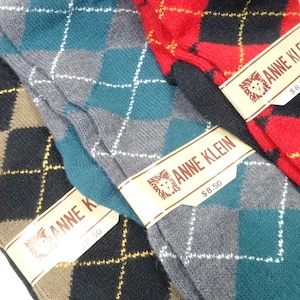 May include: Three pairs of Anne Klein socks featuring an argyle pattern. One pair is tan and black, one is teal and grey, and one is red and black. Each pair has a paper band with the text ANNE KLEIN and a lion logo printed on the front.