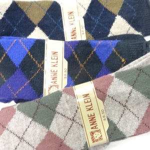 May include: Three pairs of Anne Klein socks featuring an argyle pattern. The top pair is navy, olive, and white. The middle pair is blue, black, and purple. The bottom pair is grey, green, and mauve. Each pair has a paper band with the text Anne Klein.