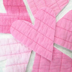 May include: A collection of pink heart-shaped fabric pieces with stitched lines, arranged on a light blue floral patterned background. The hearts vary in shades of pink, creating a layered effect.