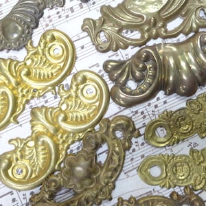 May include: A collection of assorted brass furniture hardware pieces, including ornate drawer pulls, decorative corner plates, and rectangular escutcheons, all featuring intricate embossed floral and scrollwork patterns with varying metallic finishes.