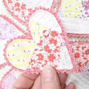 May include: A handmade, heart-shaped patchwork item with a pink scalloped edge. The hearts are made of various floral and patterned fabrics in red, yellow, purple, and blue. The item is likely a decorative piece or part of a larger craft project.