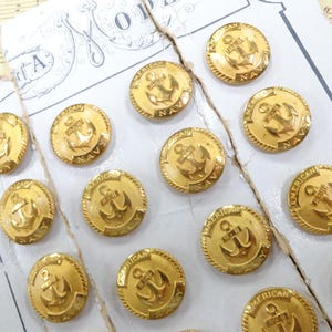May include: A set of twelve round gold-toned metal buttons arranged on a white card. Each button features a raised anchor emblem in the center with the text AMERICAN NAVY embossed along the circular border. The buttons have a textured edge.