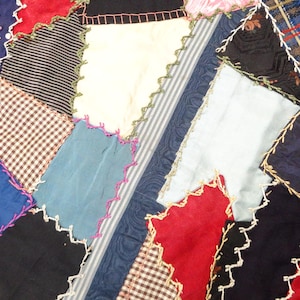 May include: Close-up of a patchwork textile with various fabric patterns and colors. The quilt features red, blue, black, white, and patterned fabrics, stitched together with decorative zigzag edges. The overall design is a mix of geometric shapes.