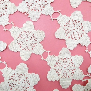 May include: Close-up of a series of white, crocheted floral doilies. Each doily is connected to the next, forming a continuous garland. The delicate, openwork design is set against a pink background.