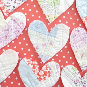 May include: A collection of handmade fabric heart decorations in various floral patterns and colors, including pink, purple, and blue. The hearts are arranged on a red background with white polka dots, creating a charming and festive display.