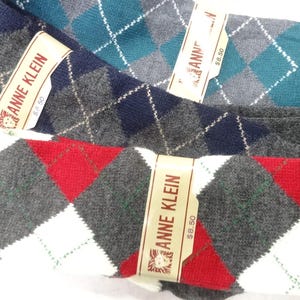 May include: Three pairs of Anne Klein socks featuring an argyle pattern. One pair is teal and grey, one is navy and grey, and one is red, white, and grey. Each pair is secured with a paper band displaying the Anne Klein brand name and logo.