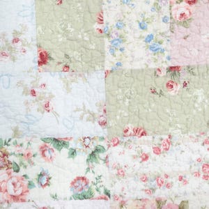 May include: A quilted fabric featuring a patchwork design of rectangular panels. The pattern displays various floral motifs in shades of pink, sage green, and light blue on white backgrounds, with a visible stitched texture across the entire surface.