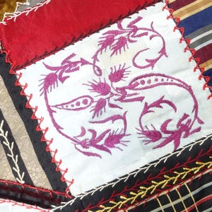 May include: Close-up of a patchwork textile with various fabric patterns and colors. A white square features a magenta floral design. Other fabrics include stripes, plaids, and solids in red, black, and gold. The pieces are stitched together with contrasting thread.