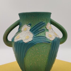 May include: A green ceramic vase with two handles, featuring white and yellow floral designs. The vase has a textured, speckled finish and a wide opening. The handles and floral details are a darker shade of green.