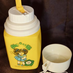 Puede incluir: Termo amarillo con tapa blanca y un personaje de Cabbage Patch Kids en la parte delantera. El termo tiene una taza blanca con asa.
