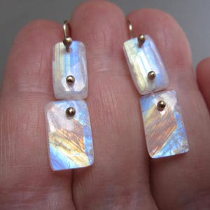 May include: A pair of rectangular earrings with iridescent, multi-colored stones. Each earring has a gold-colored hook and two gold-colored studs. The stones display hues of blue, yellow, and orange, creating a shimmering effect. The earrings are displayed against a neutral background.