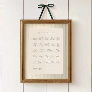 May include: A framed print of "THE CURSIVE ALPHABET" in a gold frame, hanging from a dark green ribbon. The print features the cursive alphabet in black ink on a cream-colored background with vertical stripes.