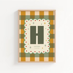 Puede incluir: Un estampado enmarcado con un fondo a cuadros verde y amarillo. El estampado tiene un borde decorativo con puntos rojos y estrellas, que enmarca una gran letra "H" verde. El nombre "Hayden" y una fecha est&aacute;n impresos debajo.