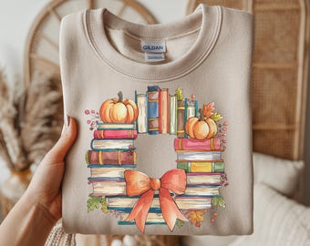 May include: Beige sweatshirt with a watercolor design of a wreath made of books, pumpkins, and a bow. The wreath is composed of stacked books in various colors, with two orange pumpkins on top. The bow is a burnt orange color.