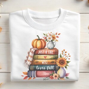 May include: White sweatshirt featuring a stack of books with the text "Just a Girl Who Loves Fall." A pumpkin, vase of flowers, and sunflower are on top. The design has fall colors.