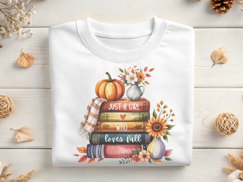 May include: White sweatshirt featuring a stack of books with the text "Just a Girl Who Loves Fall." A pumpkin, vase of flowers, and sunflower are on top. The design has fall colors.