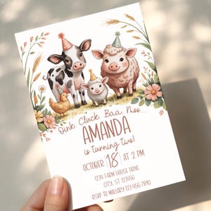Pode incluir: Convite de aniversário em aquarela com uma vaca, uma ovelha e um cordeiro usando chapéus de festa. O convite inclui o texto "Amanda está fazendo dois anos!" e detalhes do evento. Acentos florais e de trigo emolduram a ilustração dos animais da fazenda.
