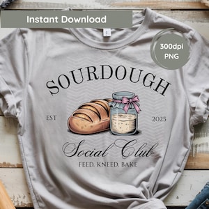 Può includere: T-shirt grigio chiaro con un design grafico. Il design include la parola "SOURDOUGH", una pagnotta di pane e un barattolo di pasta madre. È presente anche il testo "Social Club" e "Feed. Knead. Bake".