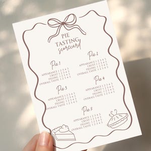 May include: A cream-colored pie tasting scorecard with a brown border and text. The card features a bow at the top and sections for rating appearance, crust, filling, and overall taste for five pies. Includes pie slice and whole pie illustrations.