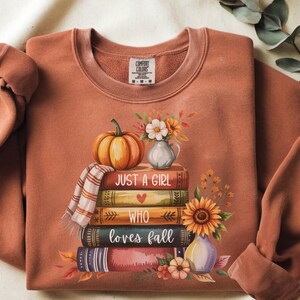 May include: Rust-colored sweatshirt with a fall-themed graphic. A stack of books is topped with a pumpkin, scarf, and flowers. The text "JUST A GIRL WHO LOVES FALL" is printed on the books. A sunflower and vase are also included.