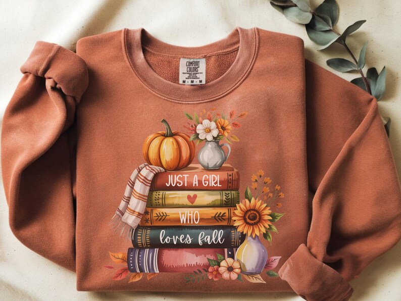 May include: Rust-colored sweatshirt with a fall-themed graphic. A stack of books is topped with a pumpkin, scarf, and flowers. The text "JUST A GIRL WHO LOVES FALL" is printed on the books. A sunflower and vase are also included.