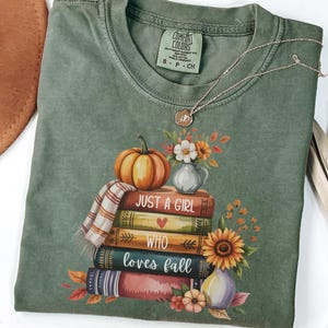 May include: Green Comfort Colors t-shirt with a fall-themed graphic. The graphic features a stack of books, a pumpkin, flowers, and the text "Just a Girl Who Loves Fall." A gold necklace with a pendant is also visible.
