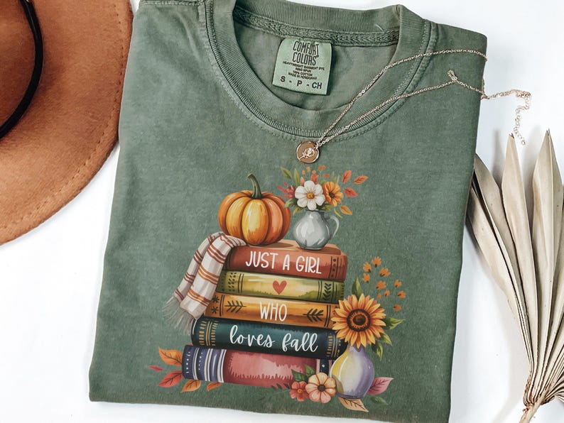 May include: Green Comfort Colors t-shirt with a fall-themed graphic. The graphic features a stack of books, a pumpkin, flowers, and the text "Just a Girl Who Loves Fall." A gold necklace with a pendant is also visible.
