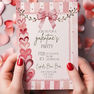 May include: A pink and white Galentine's Party invitation with a bow and heart accents. The invitation includes the text "Join us for a Galentine's Party" and event details. The invitation is held by a person with red nail polish.
