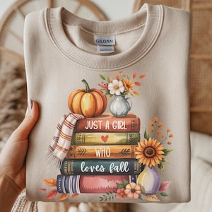May include: Beige sweatshirt with a fall-themed graphic. The design features a stack of books, a pumpkin, flowers, and the text "JUST A GIRL WHO LOVES FALL". The graphic is in warm autumn colors.