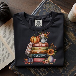 May include: Black t-shirt with a fall-themed graphic. The design features a stack of books, a pumpkin, flowers, and the text "Just a Girl Who Loves Fall." A small plaid scarf is draped over the books. A candle and a decorative pumpkin are in the background.