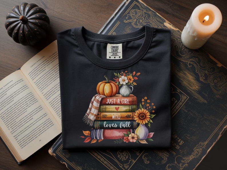 May include: Black t-shirt with a fall-themed graphic. The design features a stack of books, a pumpkin, flowers, and the text "Just a Girl Who Loves Fall." A small plaid scarf is draped over the books. A candle and a decorative pumpkin are in the background.