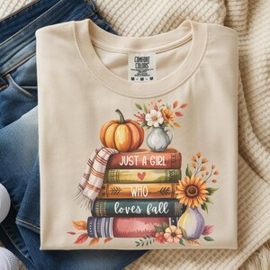 May include: Cream-colored t-shirt with a fall-themed graphic. The design features a stack of books, a pumpkin, flowers, and the text "Just a girl who loves fall." The shirt is paired with blue jeans and white sneakers.