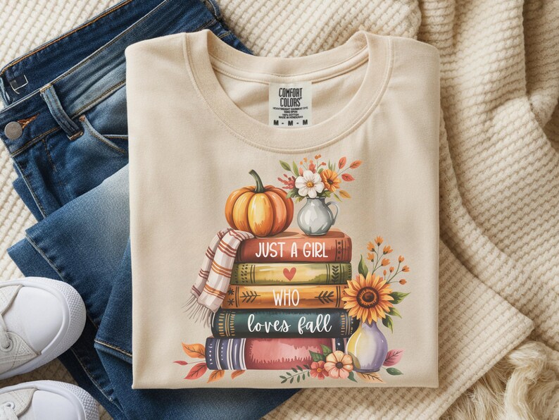 May include: Cream-colored t-shirt with a fall-themed graphic. The design features a stack of books, a pumpkin, flowers, and the text "Just a girl who loves fall." The shirt is paired with blue jeans and white sneakers.