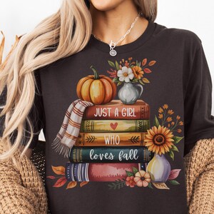 May include: Dark brown t-shirt with a fall-themed graphic. The design features a stack of books, a pumpkin, flowers, and the text "Just a Girl Who Loves Fall". The shirt has a round neckline and short sleeves.