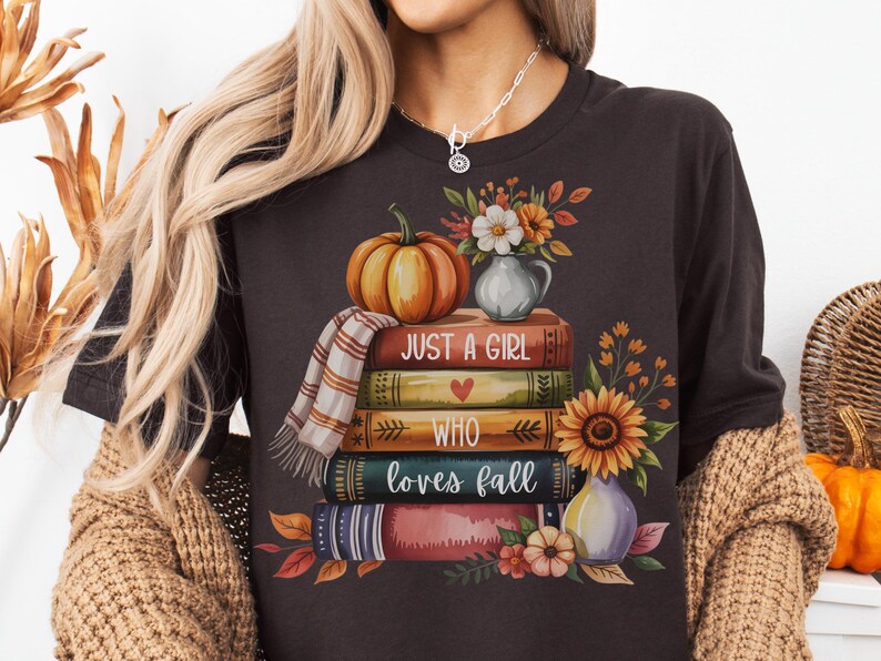May include: Dark brown t-shirt with a fall-themed graphic. The design features a stack of books, a pumpkin, flowers, and the text "Just a Girl Who Loves Fall". The shirt has a round neckline and short sleeves.