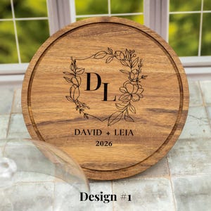 May include: A round wooden cutting board with a decorative floral wreath and the initials "DL" engraved in the center. The names "DAVID + LEIA" and the year "2026" are also engraved. The design is labeled "Design #1".