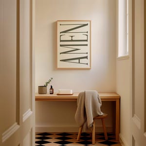 K&ouml;nnte beinhalten: Ein gerahmter Druck mit dem Wort "AMEN" in einer vertikalen, stilisierten Schriftart. Der Druck h&auml;ngt an einer wei&szlig;en Wand &uuml;ber einem Holztisch mit einer Pflanze, einer Flasche und einem wei&szlig;en rechteckigen Objekt darauf. Ein Holzstuhl mit einer grauen Decke dar&uuml;ber befindet sich im Vordergrund.