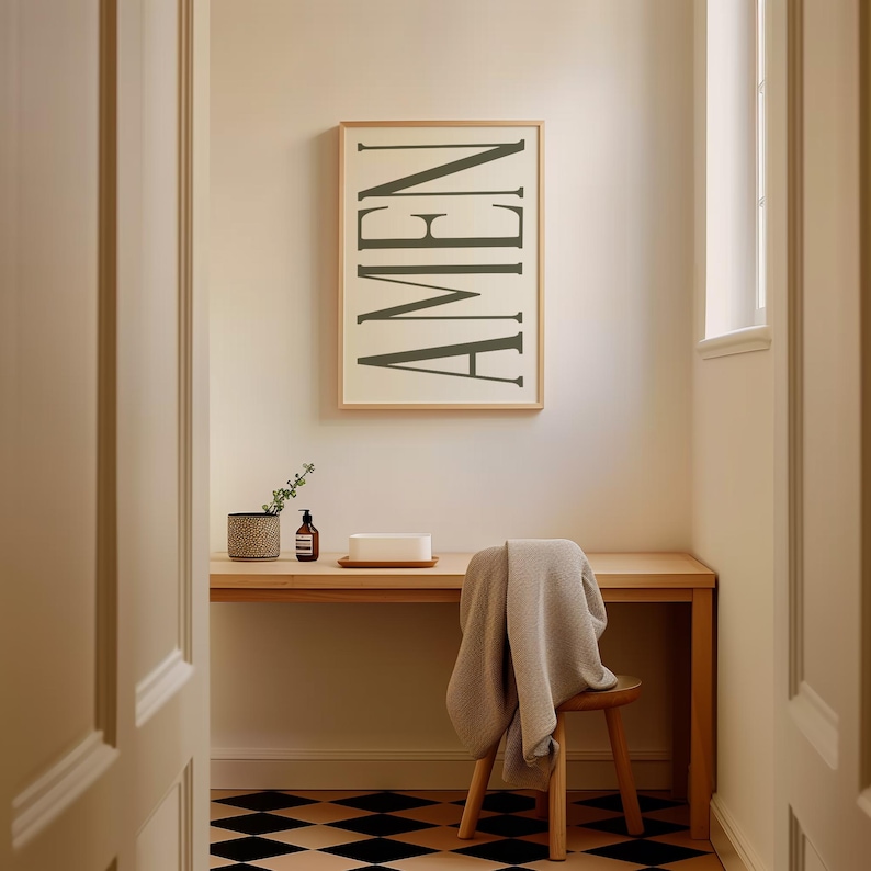 K&ouml;nnte beinhalten: Ein gerahmter Druck mit dem Wort "AMEN" in einer vertikalen, stilisierten Schriftart. Der Druck h&auml;ngt an einer wei&szlig;en Wand &uuml;ber einem Holztisch mit einer Pflanze, einer Flasche und einem wei&szlig;en rechteckigen Objekt darauf. Ein Holzstuhl mit einer grauen Decke dar&uuml;ber befindet sich im Vordergrund.
