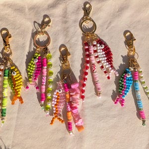 May include: Five beaded fish keychains in various colors, including yellow, green, pink, red, and blue. Each keychain features a gold-colored clasp and a beaded tail. The fish designs are detailed with colorful beads.
