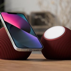Könnte beinhalten: Ein modernes, weinrotes Telefonständer- und Lautsprecher-Set. Der Telefonständer hält ein Smartphone mit farbenfrohem Display. Der Lautsprecher hat eine weiße Oberseite. Beide Artikel haben ein geripptes Design und stehen auf einer Holzoberfläche.