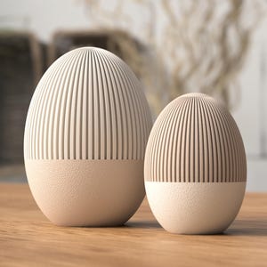 Könnte beinhalten: Zwei eiförmige Dekorationsgegenstände. Der obere Teil jedes Objekts hat vertikale Linien, während der untere Teil eine strukturierte Oberfläche hat. Die Objekte sind beige und stehen auf einer Holzoberfläche. Das größere Objekt ist etwa 18 cm hoch.