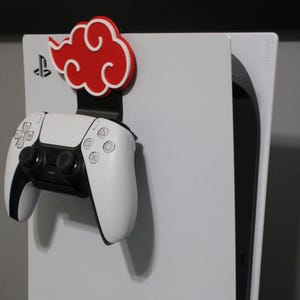 May include: A white PlayStation 5 console with a red cloud-shaped controller holder attached to the side. A white PlayStation 5 DualSense controller is hanging from the holder.