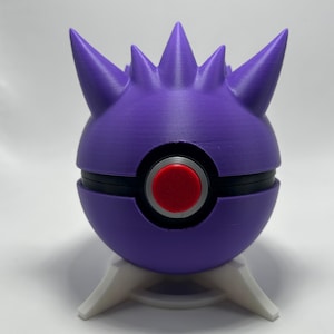May include: A purple and black Poke Ball with a red center button and white stand. The top of the ball has several purple spikes. The ball is a 3D-printed item.